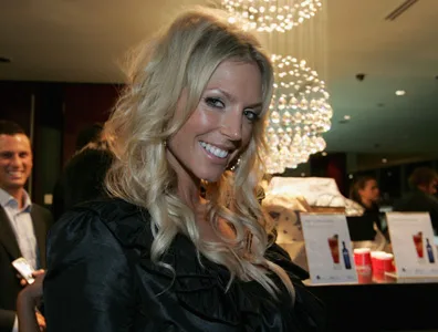 A woman with long blonde hair smiles at a party, with drinks and people in the background.