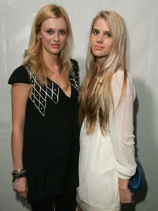 Two women with long hair pose together; one in a black dress and the other in a white dress, against a neutral background.