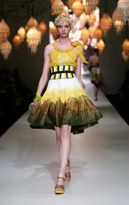 Model wearing a yellow gradient dress with a tied shoulder strap walks runway lit by hanging lanterns.