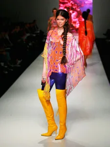 Model in colorful outfit with a vibrant top, purple tights, and yellow boots walking on a runway at Australian Fashion Week.