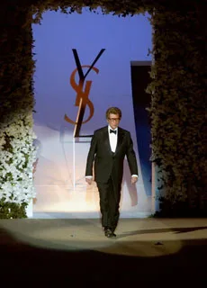 Yves Saint Laurent walking on a runway in a tuxedo with the YSL logo backdrop, surrounded by floral arrangements.