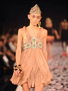 Model in beige tribal dress and hat walks on runway.