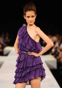 Model walking runway in a purple, one-shoulder, ruffled dress, with hands on hips during a fashion show.