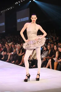 A model walks the runway in a beige, ruffled dress and white leggings at a fashion show, with audience in the background.