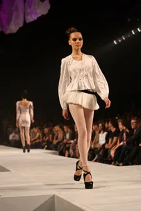 Model walking on runway in white top and leggings, wearing black heels, with audience watching.