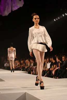 Model walking on runway in white top and leggings, wearing black heels, with audience watching.