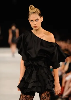 Model on runway wearing a one-shoulder black dress with ruffles and unique updo hairstyle at L'Or&eacute;al Paris fashion show.