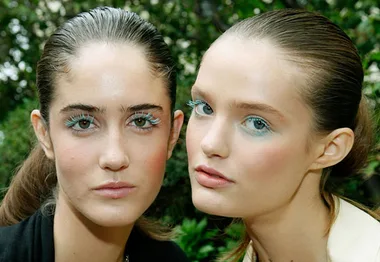 Two models with slicked-back hair and bright blue eye makeup pose outdoors.