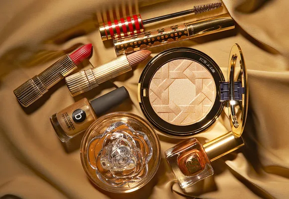 Luxury makeup products, including gold compacts, lipsticks, and a perfume bottle, arranged on a silky fabric.