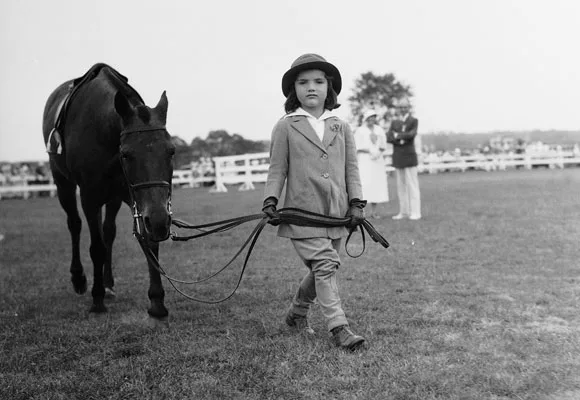 Young girl in riding attire leads a horse on a grassy field, with fenced spectators and trees in the background.