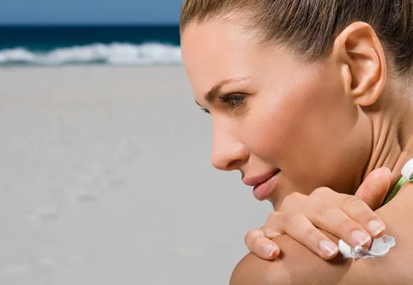 A woman applies sunscreen to her shoulder on a sunny beach with the ocean in the background.