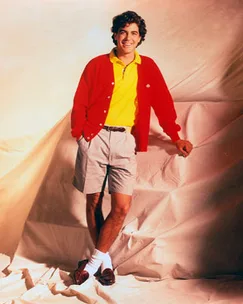 Man in bright clothing stands with one leg crossed, wearing a red cardigan, yellow shirt, and gray shorts on a draped fabric background.