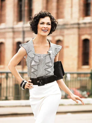 A woman in a ruffled top and white skirt smiling outdoors, with a city background.