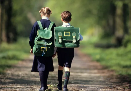 Two children in uniforms walking with green backpacks on a path surrounded by trees.
