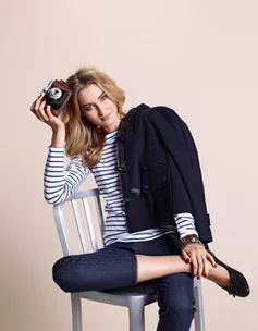 A woman in a striped shirt sits on a chair, holding a vintage camera, with a coat draped over her shoulder.