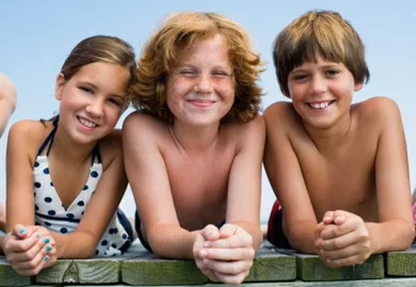 Three smiling children lying on a wooden dock on a sunny day, with a clear blue sky in the background.