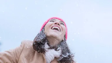 Woman in winter clothes joyfully looking up as snow falls around her.