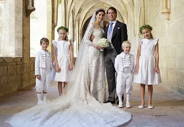 Royal wedding with the couple and children in elegant attire, floral crowns adorning the girls, in a historic stone corridor.