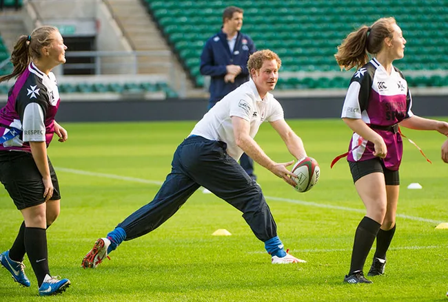 Coach in white shirt passes rugby ball to schoolgirls during a practice session on a green field.