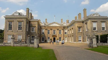 Grand, historic mansion with classical architecture, featuring columns and chimneys; visitors walking on the gravel path.