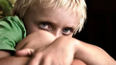 Young child with blonde hair gazing intently, partly hiding face with arms, in thoughtful pose.