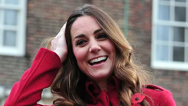 A woman in a red coat smiles while touching her hair, with a brick building in the background.