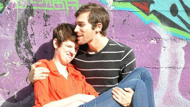 A couple sits affectionately in front of a colorful graffiti wall, with the man holding the woman in his arms.