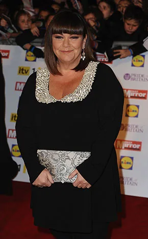 A person in a black outfit with a jeweled neckline holds a clutch on a red carpet event with people and logos in the background.