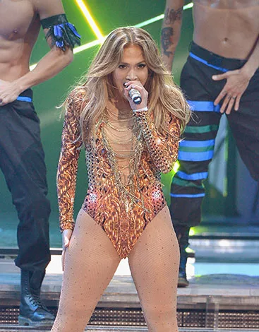 Singer in a sparkly bodysuit performs energetically with backup dancers on stage.