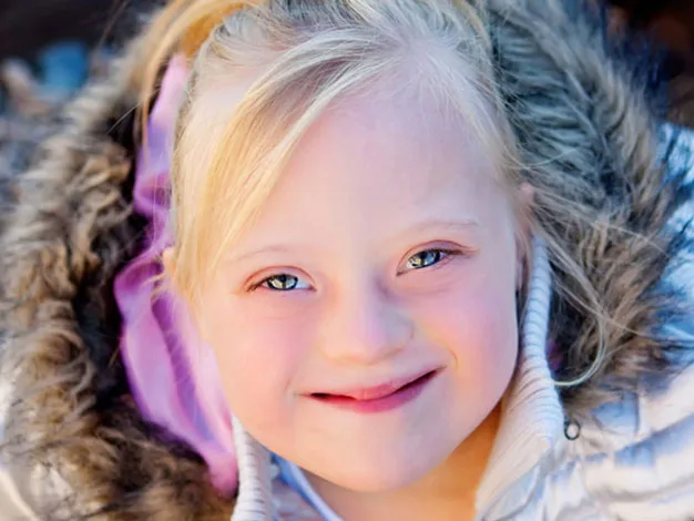 Young girl with blonde hair, wearing a coat with a fur-lined hood, smiles warmly at the camera.