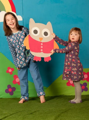 Two children smiling, holding a large paper owl cutout in front of a colorful backdrop with flowers and a rainbow.