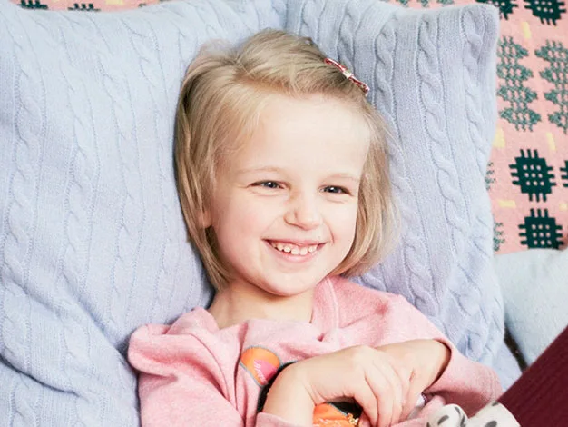 Young child smiling, lying on blue knitted pillows, wearing a pink sweatshirt.