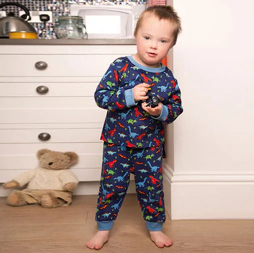 A young child in dinosaur pajamas stands by a kitchen counter holding a toy, with a teddy bear nearby.