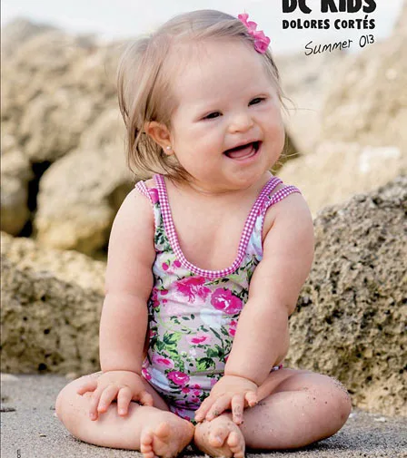 A smiling toddler wearing a floral swimsuit with a pink flower clip sits on a rocky beach.
