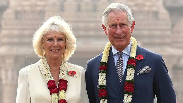 Royal couple wearing garlands, smiling outdoors with stone architecture background.