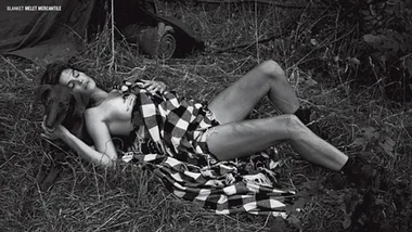 A woman lies in a field, partially covered with a checkered blanket, next to a dog in a serene pose.