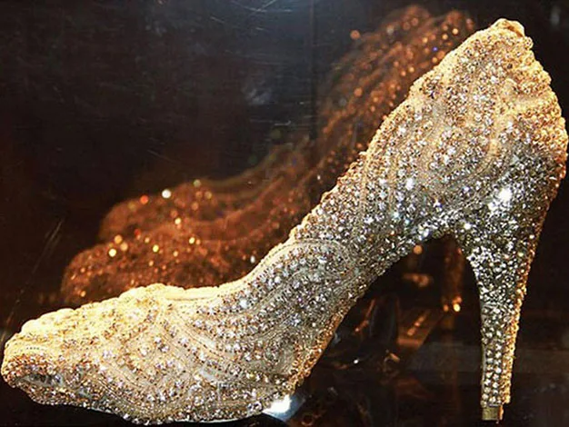 Golden high-heeled shoe encrusted with sparkling crystals, displayed on a reflective surface.