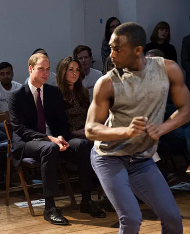 Prince William and Kate Middleton watch a dance performance by a man in a sleeveless shirt during an event.