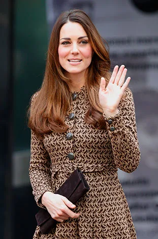 A person in a patterned dress waves, holding a clutch with long brown hair, outdoors.