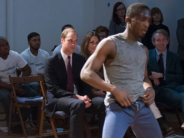 Audience watches dancer perform; man in focus wears gray tank top, blue pants, surrounded by seated spectators.