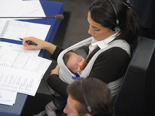 Woman in a meeting holding a baby in a sling, wearing headphones, reviewing documents.