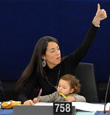 Woman with child raises thumb in approval at a conference table, seated with papers and toys numbered 758.