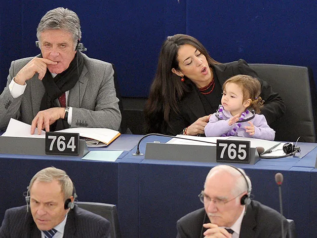 Man in suit listens, woman holds baby at conference table, numbered seats 764-765, others in background.