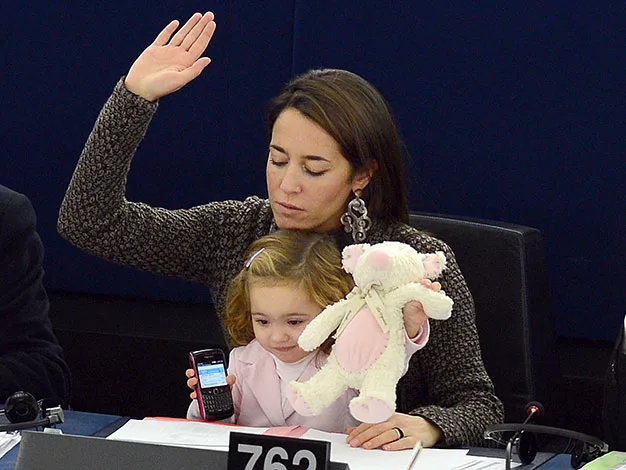 A woman raises her hand in a meeting while holding a child with a toy and phone.