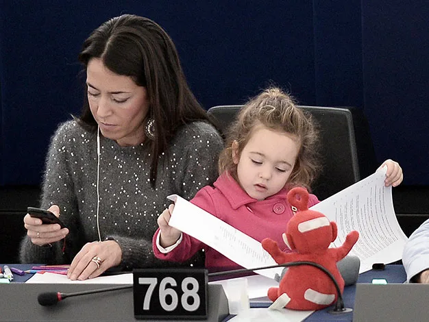Woman and child at a table; woman using a phone, child holding papers, with a red toy in front.