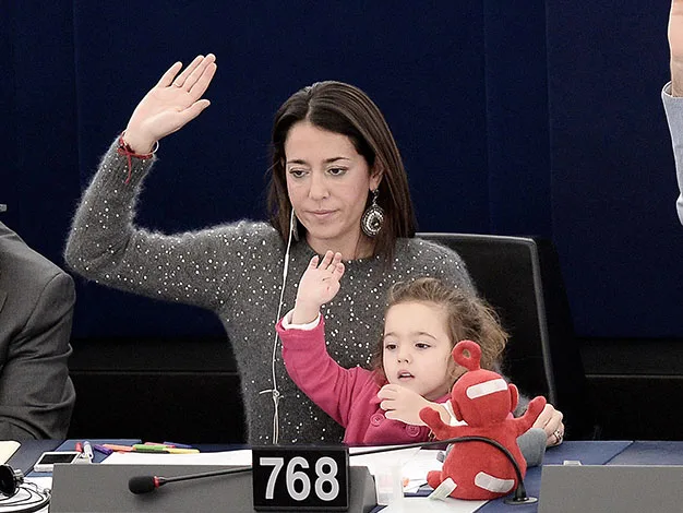 Woman and child raising hands at desk with toy; seated in an official setting, number 768 visible.