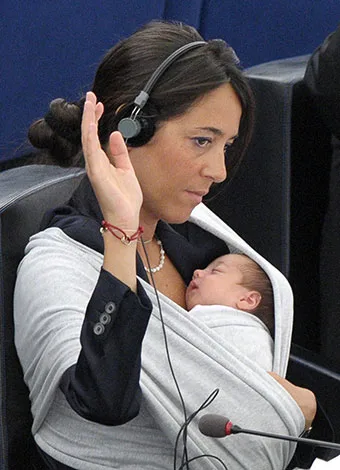 A woman wearing a headset in a formal setting, holding a sleeping baby in a sling, raising her hand during a session.