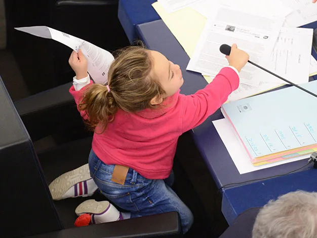 Child in pink sweater and jeans holding papers, reaching for a microphone at a desk cluttered with documents.