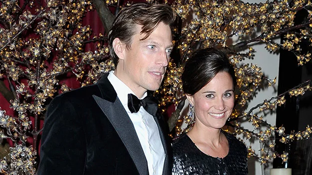 Two people in formal attire smiling at an event with decorative lighting and floral background.