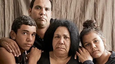 A family of four, somber expressions, gather closely in front of a neutral background.